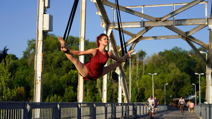 Circus performer on aerial straps performs an aerial act at dawn. flexible, athletic, aerial gymnast in city on metal trusses of bridge engaged in aerial gymnastics. urban acrobatic street show. split