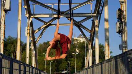 Beautiful young woman gymnast hanging in a flexible pose on an iron bridge in the rays of sunlight Professional circus performer, graceful flying on aerial straps