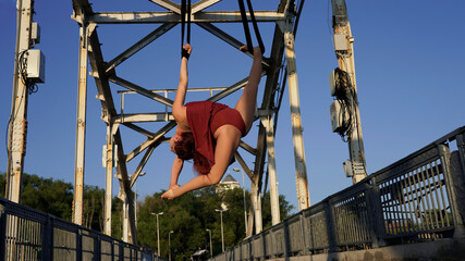 Beautiful young woman gymnast hanging in a flexible pose on an iron bridge in the rays of sunlight Professional circus performer, graceful flying on aerial straps