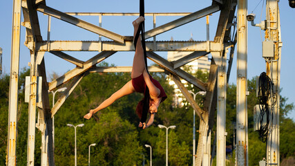 Beautiful young woman gymnast hanging in a flexible pose on an iron bridge in the rays of sunlight Professional circus performer, graceful flying on aerial straps