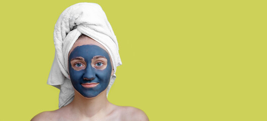 A young teenage girl in a white towel on her head, with a cosmetic clay mask on her face, bare shoulders on a yellow isolated background.The concept of Spa treatments, Facials. Banner, copy space