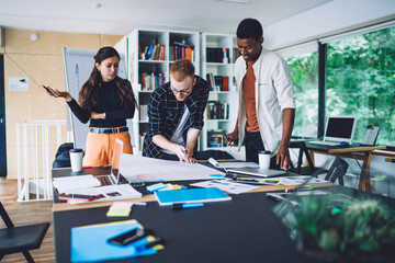 Group of smart casual developers working with project sketches for architect order, young colleagues collaborating at desktop table on blueprint illustrations discussing ideas and communicate
