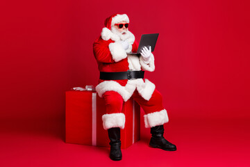 Full length photo of retired old man white beard hold netbook sit box take curious child present order wear santa x-mas costume sunglass headwear leather boot isolated red color background