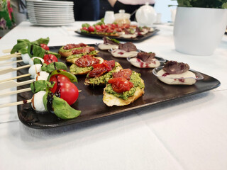 black tray with snacks on a white tablecloth on the table. On a tray canapes with cherry tomatoes, tofu, herbs and meat. Holiday concept