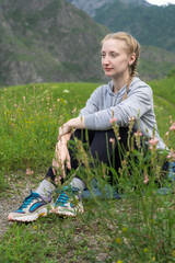 Young woman hiker in sportswear sits resting on a green meadow against the backdrop of a mountain. Summer, travel, vacation or relaxation concept.
