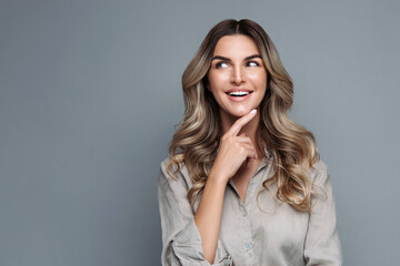 Fototapeta na wymiar Blonde woman with curly hair dressed in linen shirt, smiles pleasantly, looking at blank space, happy with discounts and sales, arm on chin, isolated on grey background. Advertising service, low price