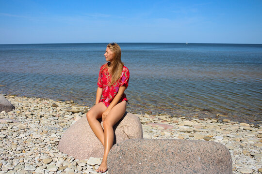 A beautiful, long-legged blonde girl with loose hair in a red shirt sits on the beach on a large stone and looks away.