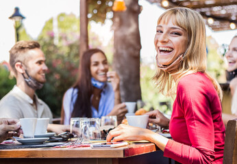 Happy young woman wearing face mask smiling at the camera at the restaurant cafè. Group of friends drinking coffee sitting at the bar.