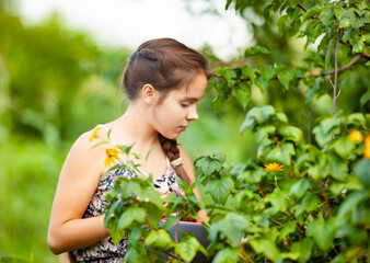 Side view of smiling young girl with long brown braid sitting on wooden chair while collecting harvest in garden. Female teenager enjoying picking delicious berries, holding bowl.