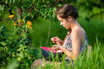 Side view of young girl with brown braid sitting on wooden chair while collecting harvest in garden. Female teenager with eyes closed enjoying eating delicious berries, holding bowl.