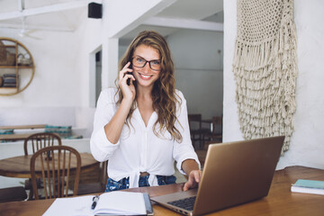 Obraz premium Cheerful student looking at camera while phoning and using laptop in cafe