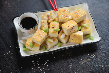 Chunks of pan fried tofu cheese with green onion, sesame and soy sauce, studio shot on a dark brown stone background