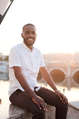 Fototapeta na wymiar Portrait of smiling young bearded African-American guy in white shirt sitting on marble railing against city river