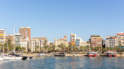 Landscape of the port of alicante with the promenade full of palm trees and the sea with many boats with a clear blue sky in alicante, spain