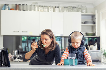 Woman in the kitchen eating and watching at electronic tablet. Child sits on table in headphones and watch at mobile phone