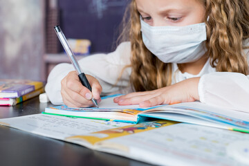 Child with face mask back at school after covid-19 quarantine and lockdown. Young elementary school girl with face protective mask writing in a book. Focus on pen. education concept.