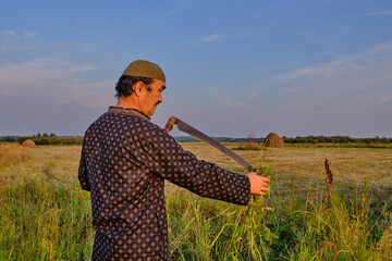 An senior asian man in a skullcap and traditional clothes cleans a hand scythe with grass in a...