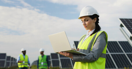 Portrait of electrician engineer in safety helmet and uniform using laptop checking solar panels. Female technician at solar station.