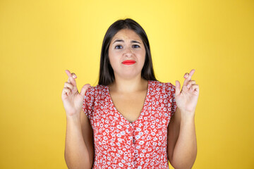 Young beautiful woman over isolated yellow background gesturing finger crossed smiling with hope and looking side