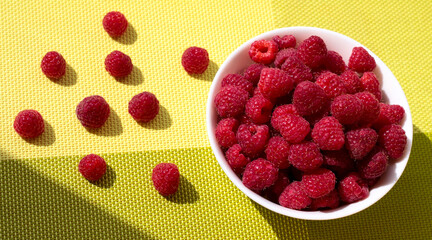 Raspberries in a white bowl on a green background. Ripe berries are scattered.