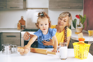 A mother and her little daughter prepare ingredients for baking. On the table are eggs, milk, flour, and a rolling pin. A little girl reaches for an egg in a glass bowl. Family vacation, time together
