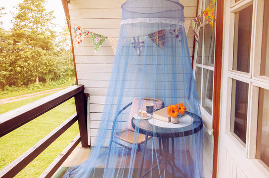 Table set on country home balcony, protected by mosquito net, to keep away annoying bugs. Book, cake, mug, bouquet of flower on table. 