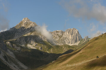 landscape of the mountain peaks of the Alps, rugged and rocky mountains, part of the French Alps mountain range. The sky between clear blue, is covered with white clouds, the valley and grass