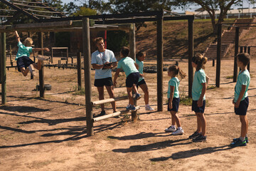 Male fitness coach instructing while kids hanging on monkey bars at a boot camp