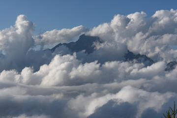 Naklejka na ściany i meble Rocky mountain peak of the Alps, emerging from the clouds, seen above the clouds, from within the cloud. Above all, the sky is blue and the clouds are below the peaks and the photographer, photo taken