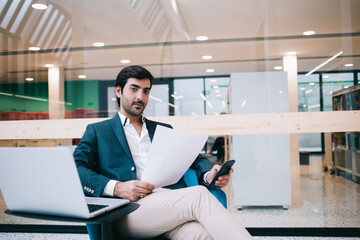 Naklejka na ściany i meble Portrait of South Asian businessman sitting at desktop with modern laptop computer in office interior and looking at camera during time for work with financial documents, technology marketing