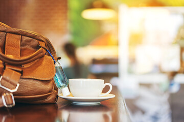 Mini alcohol gel bottle to kill Corona Virus(Covid-19) hang on a brown leather shoulder bag with hot coffee on table in cafe.New normal lifestyle.Health care concept.Selective focus on alcohol gel