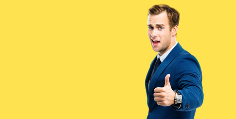 Excited happy businessman showing thumbs up like hand sign gesture, blue confident suit, on yellow color background. Handsome happy man. Copy space for some slogan or advertising text.