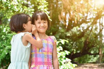 Two little sister girls whisper in ear at park outdoors. Friendship, happiness and people concept