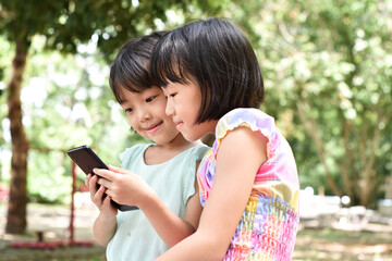 Two smiling little girl holding smartphone is watching online video in park outdoor.