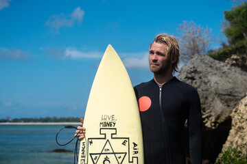 Portrait of handsome man surfer in black wetsuit, holding white surf board  and blue ocean, cliff,...