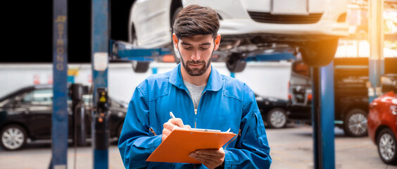 Mechanic in blue work wear uniform checks the vehicle maintenance checklist with blur lifted car in the background. Automobile repairing service, Professional occupation.