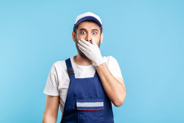Fototapeta na wymiar I won't say anyone! Scared handyman in overalls and gloves covering mouth with hand, afraid to talk, looking intimidated and frightened. Profession of service industry, courier delivery. isolated