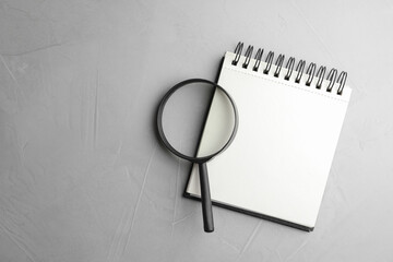 Top view of magnifier glass and empty notebook on light grey stone background, space for text. Find keywords concept