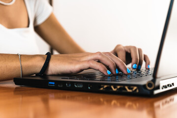 Detail of girl's hands typing on a black laptop on a wooden table and white background. Her nails are painted blue.