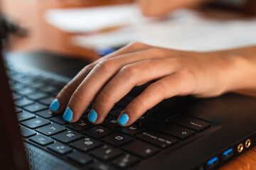 Detail of girl's hand typing on a black laptop on a wooden table. Her nails are painted blue.