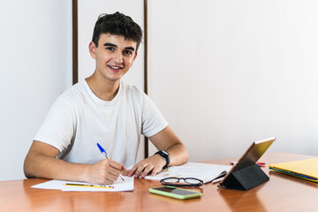 Young brunette smiling student boy writing on a paper on a wooden table with a book, glasses and pens