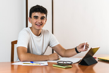 Young brunette smiling student boy using a tablet with a digital pen on a wooden table with a book, some glasses, pens and sheets of paper.