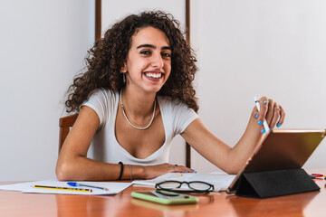 Young smiling student girl with curly brunette hair using a tablet with a digital pen on a wooden table with a book, glasses, pens and sheets of paper.