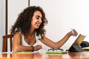 Young smiling student girl with curly brunette hair using a tablet with a digital pen on a wooden table with a book, glasses, pens and sheets of paper.