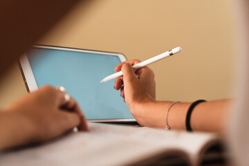 Detail of student girl hand picking up a digital pen using a tablet at a table with books and pens