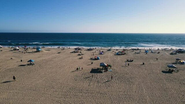 A medium paced Aerial Dolly Forward Runout from the boardwalk to the water at Huntington Beach, California over the crowds relaxing and playing at the beach around sunset.