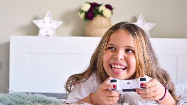 A little girl is playing a computer console on a sofa in a bright room. Concurrent games for children. Joystick for control in hands. Emotions and fun of the child. Copy space. High quality 4k footage