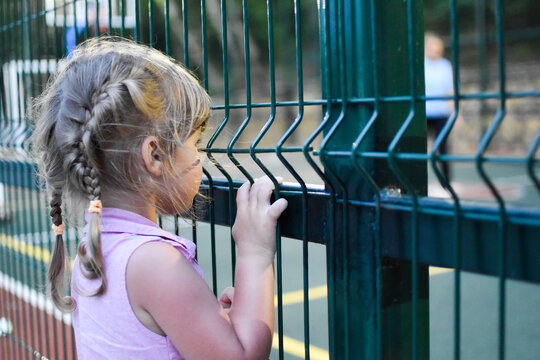 Little Child Girl Looks Behind A Metal Grate Fence. A Girl In An Orphanage