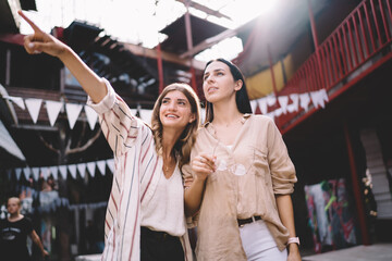 Youthful Caucasian women in stylish clothing enjoying together vacations for exploring world discussing city landscape during travel sightseeing, happy friends 20s socialising at urban setting
