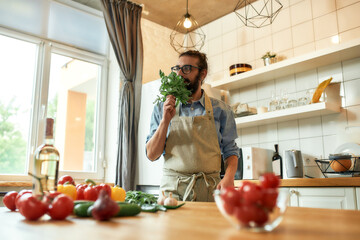 Young man, Italian cook in apron smelling basil leaves while getting ready to prepare a meal, standing in the kitchen. Cooking at home concept